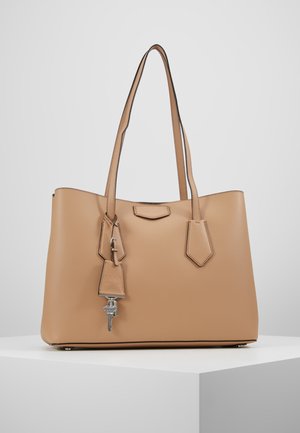 Tan leather tote bag with two shoulder straps and a small hanging leather tag holding a silver key, placed on a white pedestal.