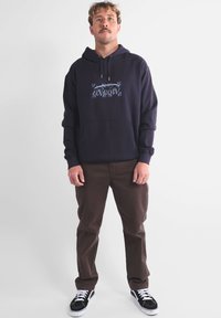 Sweat-shirt à capuche bleu marine avec un logo graphique bleu sur la poitrine, associé à un pantalon marron et des baskets noires avec des accents blancs.