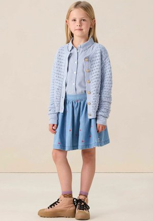 Lichtblauwe gebreide cardigan, wit en blauw gestreept shirt, spijkerrok met rode hartpatronen en beige hoge sneakers.