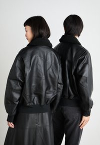 Μαύρα δερμάτινα μπουφάν bomber με μαύρο γούνα στην κολλέτα, διαθέτοντας ραμμένα μανίκια και στρίφωμα, απεικονιζόμενα από πίσω σε δ ύο άτομα.