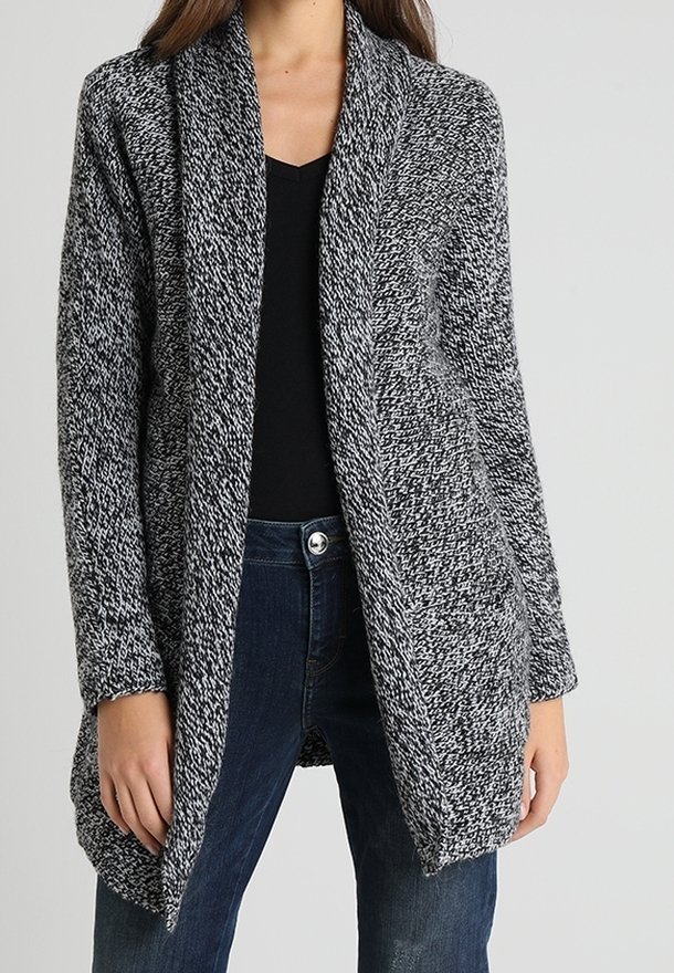Femme portant un cardigan en maille chinée noir et blanc à ouverture devant, sur un top noir et un jean bleu, debout devant un fond uni.
