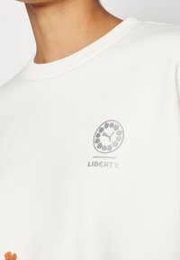 Gros plan d'une personne portant un haut blanc avec un logo floral circulaire et le mot « LIBERTY » imprimé sur la poitrine.