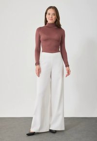 Moteris stovi prieš baltą sieną, vilkinti alyvinės spalvos golfo tipo megztinį, plačius baltus kelnių ir juodus batus, su neutralia išraiška.