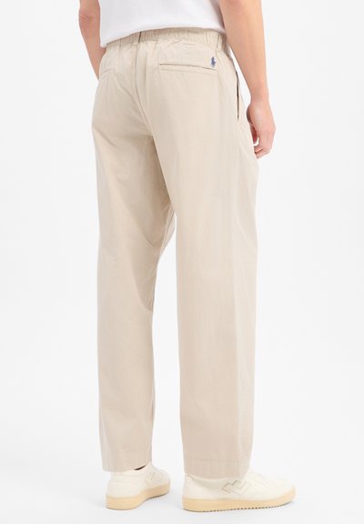 Homme portant un pantalon beige droit avec ceinture élastique et des baskets blanches, vu de dos sur fond blanc.