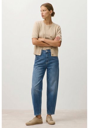Frau steht mit verschränkten Armen, trägt einen beigefarbenen Kurzarm-Pullover, eine passende Strickjacke, hoch taillierte blaue Jeans und beigefarbene Sneaker.