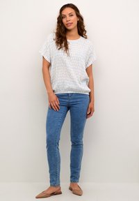 Haut à motifs bleu clair avec une coupe décontractée et des manches courtes, associé à un jean bleu slim et des ballerines beiges, sur un fond uni.