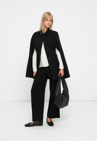 Cappotto nero con ampi bottoni, abbinato a una camicia bianca e pantaloni neri a gamba larga. La modella tiene una borsa a tracolla nera, in piedi davanti a uno sfondo bianco.