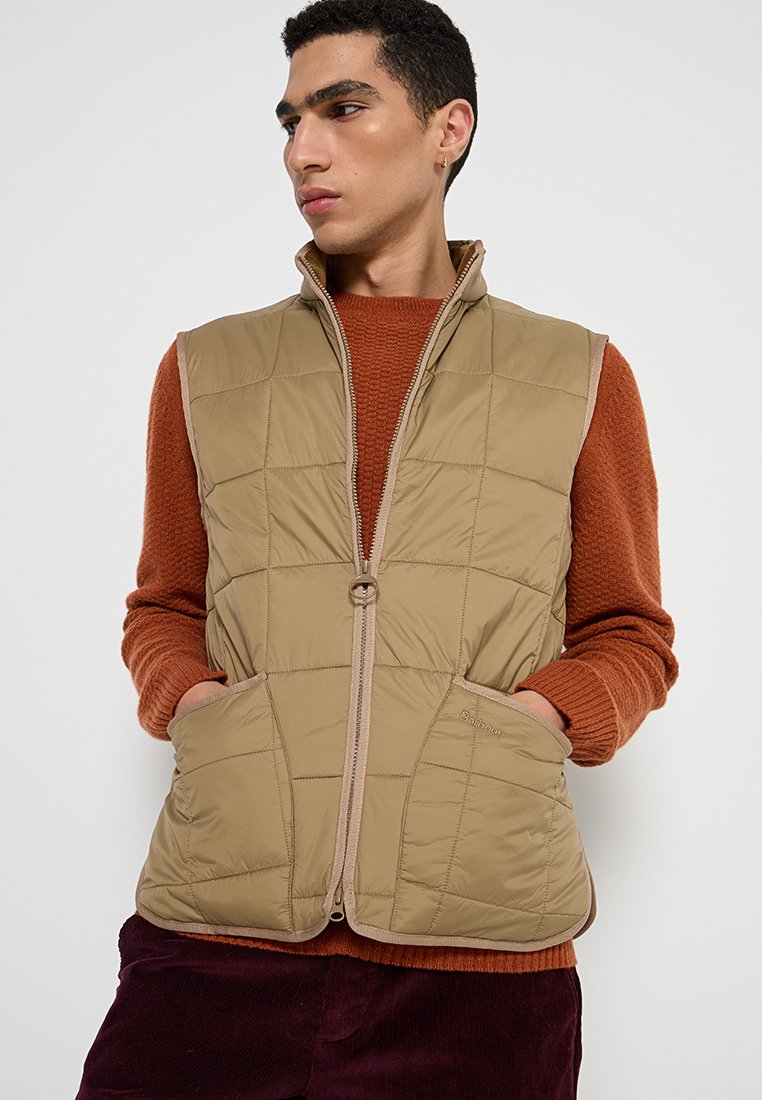 Gilet beige trapuntato con zip frontale, due grandi tasche e dettagli marroni, indossato sopra un maglione arancione, con una texture leggera e liscia.