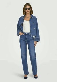 Γυναίκα φορώντας oversized μπλε τζιν μπουφάν, λευκό τοπ, ψηλόμεσα μπλε τζιν, μαύρες γόβες, γυαλιά ηλίου και αλυσίδα στον λαιμό, να στέκεται σε λευκό φόντο.