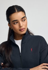 Giacca zip-up grigia scura con colletto alto, realizzata in tessuto morbido, con un piccolo logo rosso sul lato sinistro del petto. Camicia bianca sotto.