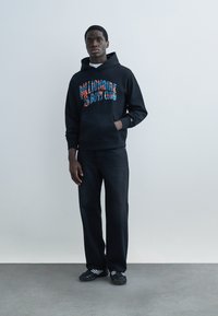Young man standing against plain wall wearing black "Billionaire Boys Club" hoodie, black pants, and black sneakers with white stripes.
