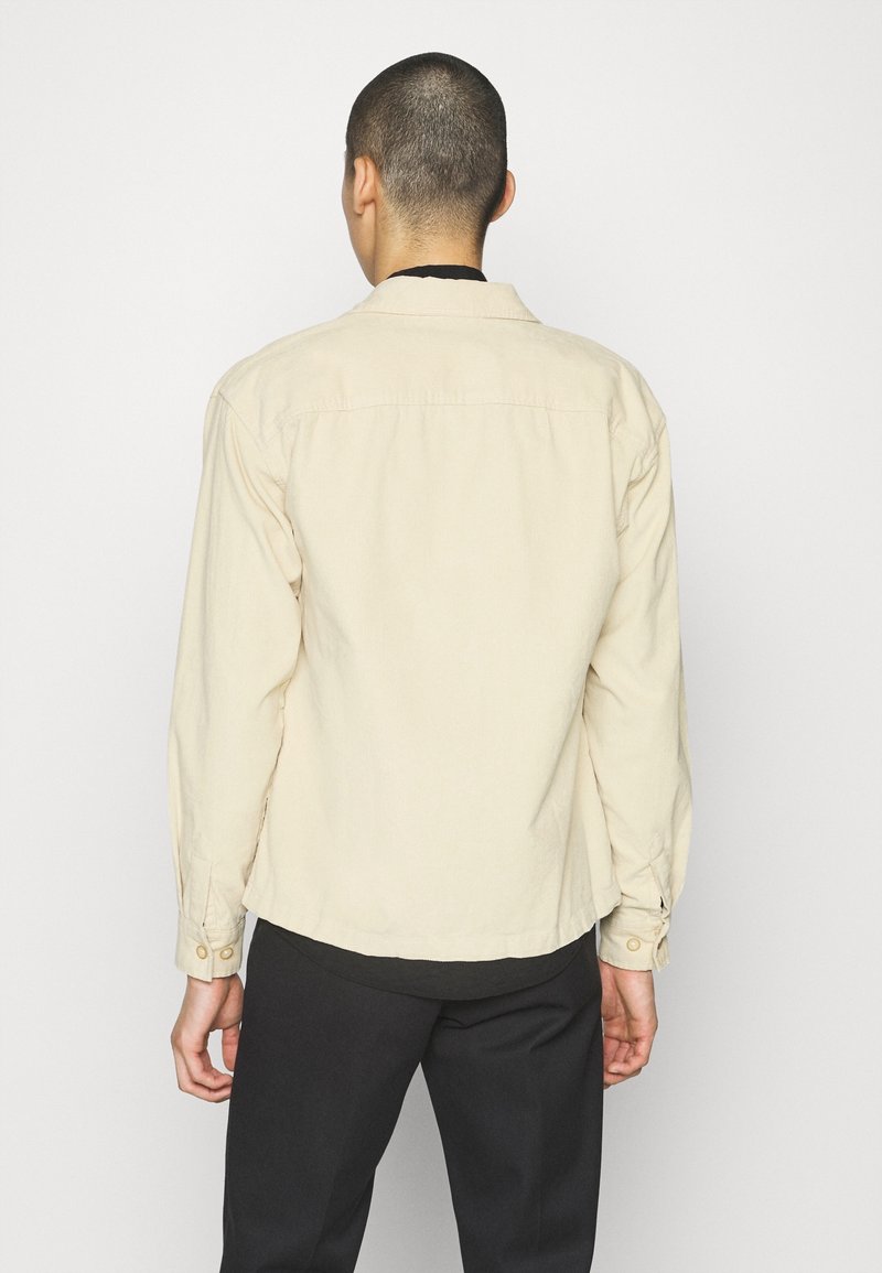 Camicia button-up beige chiaro con maniche lunghe, colletto e retro dritto. Presenta dettagli cuciti e polsini con bottoni.