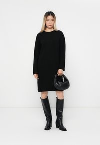 Abito in maglia oversize nero con maniche lunghe, abbinato a stivali neri lucidi fino al ginocchio e una borsa in pelle nera. Sfondo bianco semplice.