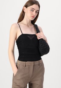 Czarna top z marszczeniami w lekkim materiale z cienkimi ramiączkami, w połączeniu z beżowymi spodniami z zamszu. Modelka trzyma torebkę z teksturowanego czarnego materiału.