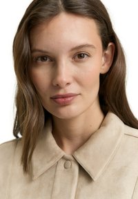 Beżowa zamszowa kurtka z ostrym kołnierzem, zapięciem na zatrzaski i gładką fakturą. Charakteryzuje się prostym designem bez widocznych wzorów ani ozdób.