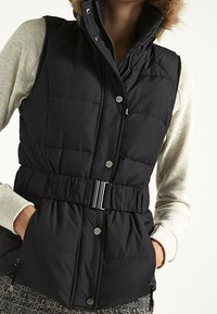 Osoba ubrana w czarną kamizelkę puchową z paskiem i kapturem wykończonym futrem, nosząca jasnobeżową koszulę z długim rękawem, z rękami w kieszeniach.