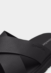 Sandale en cuir noir avec des brides croisées texturées et le logo Calvin Klein embossé sur la partie supérieure.