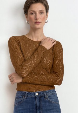 Femme aux cheveux courts châtains porte un haut en dentelle marron à manches longues et un jean bleu, debout avec une main sur l’épaule sur fond blanc.