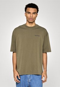 Oliwkowo zielona oversize koszulka z miękkiej bawełny, z okrągłym dekoltem i krótkimi rękawami, z subtelnym logo na lewej piersi.