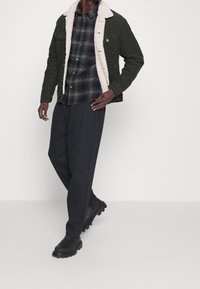 Green corduroy jacket with a cream shearling collar, black and green plaid shirt, loose dark pants, and black chunky boots.