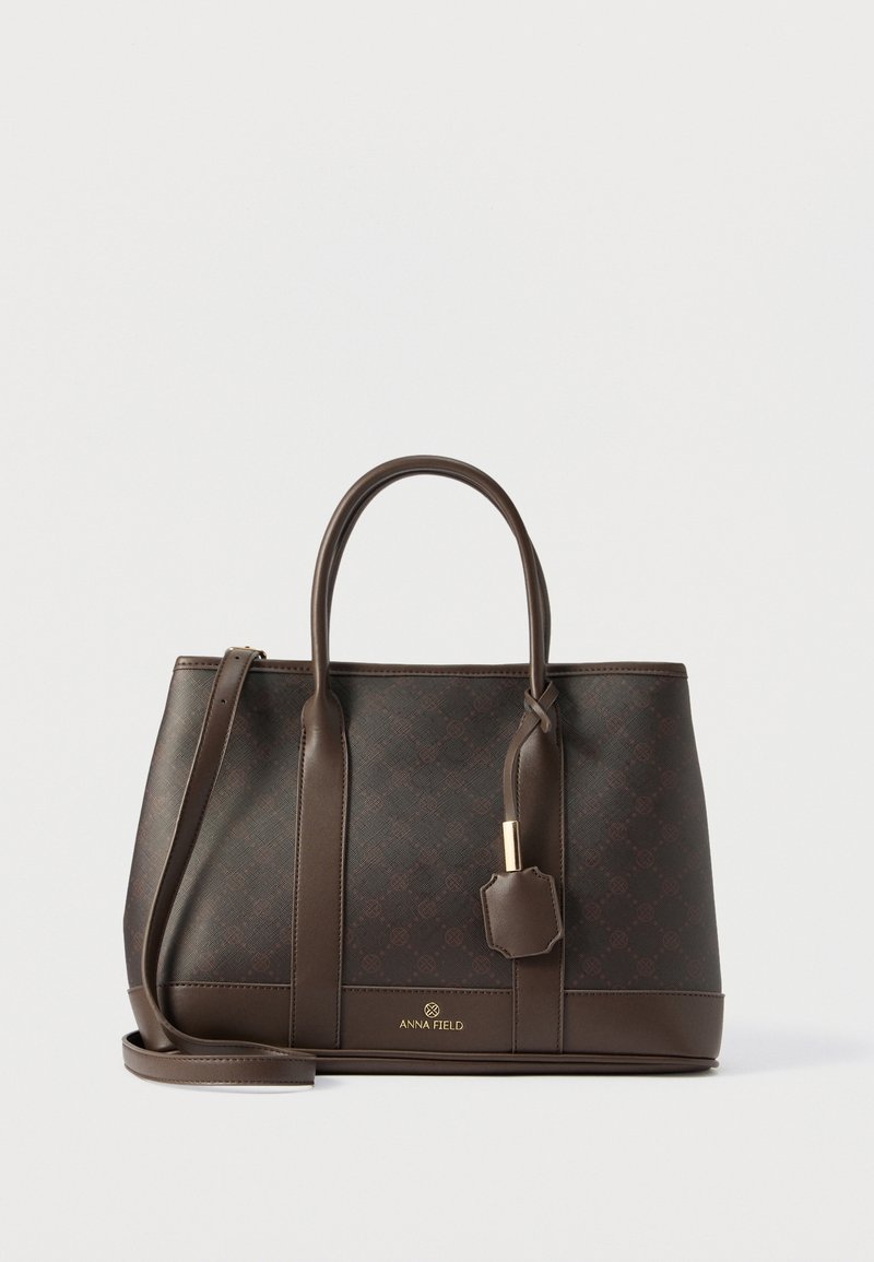 Sac fourre-tout en cuir marron foncé avec deux poignées, bandoulière amovible et petite étiquette suspendue, marqué "Anna Field" à l'avant.