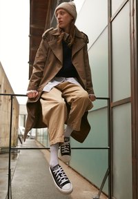 Bruine trenchcoat over een marineblauwe sweater, gestreept shirt en beige wijde broek. Zwarte sneakers met witte zolen, model zittend op een metalen reling.