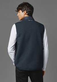 Marinblå ärmlös väst med texturerad yta, med hög krage och logodetalj på baksidan, kombinerad med en vit långärmad skjorta.