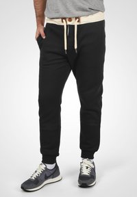 Black jogger pants with a cream waistband and drawstring, featuring side pockets and a tapered fit, paired with grey shoes.