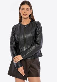 Woman wearing black zip-up leather jacket, brown leather skirt, large hoop earrings, and gold chain necklace against white background.