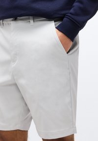 Άνδρας που φοράει ανοιχτό γκρι σορτσάκι με το χέρι στην τσέπη και ένα μπλε σκούρο μακρυμάνικο πουκάμισο που φαίνεται στη μέση και στον βραχίονα.