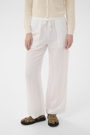 Personne portant un pantalon blanc ample à cordon, un cardigan beige en maille et des sandales marron à bout ouvert, debout devant un fond uni.