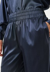 Tamsiai mėlynos elastingos juostos kelnės, turinčios lygią, blizgią tekstūrą ir laisvą siluetą. Audinys minkštai krenta, akcentuodamas subtilų blizgesį.