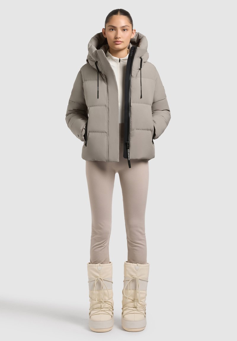 Doudoune gris clair avec capuche, fermeture éclair noire et cordons de serrage. Associée à des leggings beiges et des bottes d'hiver crème avec des accents en dentelle.