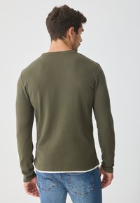 Ελιά πράσινο μακρυμάνικο πουκάμισο με ραβδωτή υφή, στρογγυλή λαιμόκοψη και ευθεία ρίγα, συνδυασμένο με τζην παντελόνι, εμφανίζοντας την πίσω όψη.