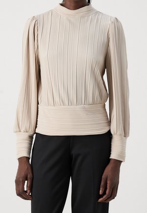 Beige plisseret bluse med lange ærmer, høj hals og tætsiddende talje båret med sorte bukser på en mørkhudet person.