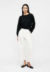 Γυναίκα που φοράει μαύρο μακρυμάνικο πουλόβερ, λευκό φαρδύ παντελόνι cropped, μαύρα φλατ παπούτσια και χρυσούς κρίκους σκουλαρίκια, στέκεται με το ένα χέρι στην τσέπη.