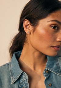 Pendientes de botón de plata con gemas claras, llevados en una oreja bronceada. El sujeto lleva una camisa de mezclilla azul claro con cuello.