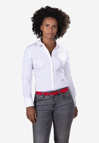 Woman with curly hair wearing a white button-up shirt, gray jeans, and a red belt, standing against a plain white background.