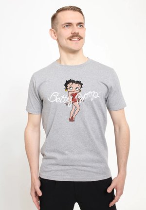 Homme avec moustache portant un t-shirt gris avec un dessin animé de Betty Boop et du texte, regardant légèrement sur le côté, debout devant un fond blanc.