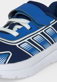 Gros plan sur une chaussure de sport bleue avec des lacets blancs, une sangle Velcro bleue, une semelle blanche et un motif latéral à rayures bleues et blanches.