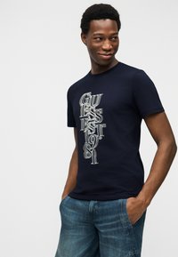 Νεαρός άνδρας με κοντά κατσαρά μαλλιά που φοράει μπλε σκούρο μπλουζάκι με λευκό κάθετο κείμενο "GUESS EST 1981" και μπλε τζιν, τα χέρια στις τσέπες.