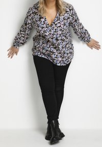 Frau, die schwarze Hosen, schwarze Stiefel und eine langärmlige Bluse mit Blumenmuster trägt, sich nach hinten lehnt und die Arme ausstreckt vor einem weißen Hintergrund.
