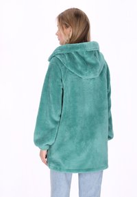 Chaqueta de forro polar azul verdoso con capucha, que presenta una textura suave, mangas largas y un ajuste holgado. El dobladillo es redondeado en la parte inferior.