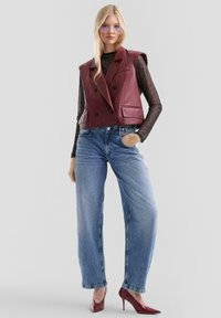 Burgunderfarbene Leder-Doppelkneifer-Weste über einem durchsichtigen schwarzen Oberteil, kombiniert mit hellblauen, hochgeschnittenen Jeans und spitzen roten High Heels.