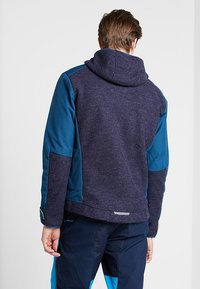 Homme portant une veste à capuche bicolore bleue et un pantalon bleu assorti, debout dos à l'appareil photo sur un fond uni.