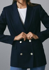 Persona che allaccia i bottoni di un blazer doppiopetto blu navy sopra una camicia bianca e jeans blu su uno sfondo grigio uniforme.