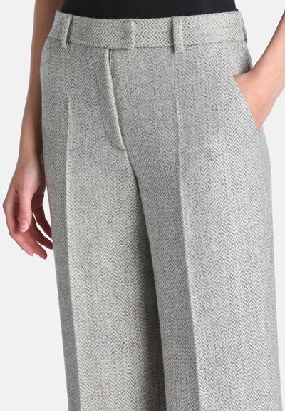 Gros plan d'une personne portant un pantalon gris à motif chevrons, avec passants de ceinture, poches latérales et une main reposant dans une poche.
