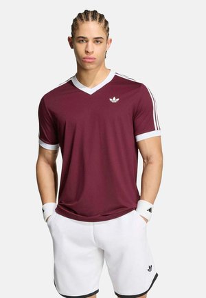 Jonge man met gevlochten haar, gekleed in een bordeauxrood Adidas sportshirt en witte korte broek met bijpassende polsbandjes, staand tegen een effen achtergrond.