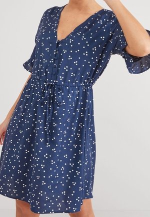 Robe bleu marine à pois blancs, col en V, manches courtes à volants et taille ajustable par cordon, avec une patte de boutonnage sur le devant.