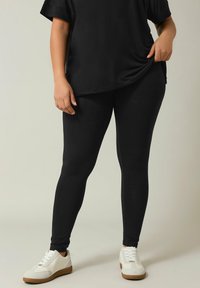 Zwarte legging en een losse zwarte top worden gedragen met witte sneakers met tan accenten. De outfit heeft een gladde, strakke textuur.